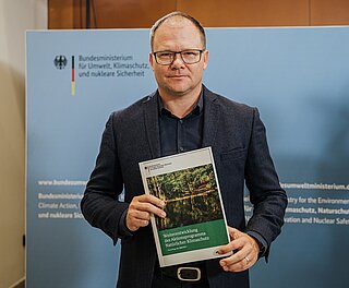 Bundesumweltminister Carsten Schneider hält die Vorschläge zur Weiterentwicklung des Aktionsprogramms Natürlicher Klimaschutzin der Hand.